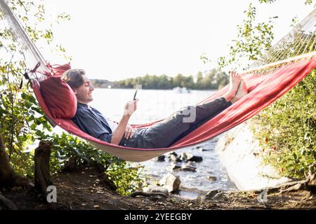 Vue latérale de l'homme souriant à l'aide d'un téléphone intelligent tout en s'allongeant dans un hamac près du lac Banque D'Images