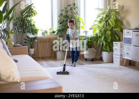 Un petit garçon européen caucasien aspire le tapis dans le salon de la maison. Banque D'Images