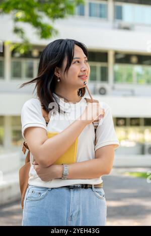 Une étudiante d'université féminine asiatique jolie et réfléchie dans des vêtements décontractés avec un sac à dos se tient sur son campus universitaire, tenant un crayon à son menton comme s. Banque D'Images
