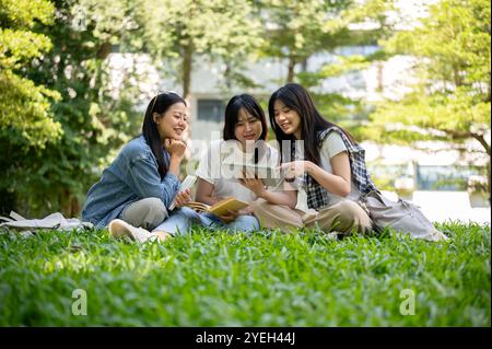 Un groupe de jolies étudiantes asiatiques s'assoit sur l'herbe dans le parc du campus de leur université, discutant d'une vidéo qu'elles regardent ensemble sur un Banque D'Images