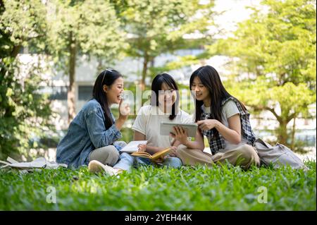 Un groupe de jolies étudiantes asiatiques s'assoit sur l'herbe dans le parc du campus de leur université, discutant d'une vidéo qu'elles regardent ensemble sur un Banque D'Images