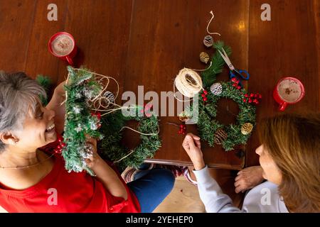 Amies féminines seniors fabriquant des couronnes de Noël à la maison, dégustant du chocolat chaud ensemble Banque D'Images