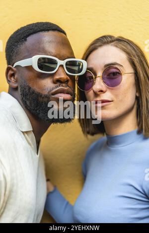 Gros plan vertical d'un couple multiracial élégant portant des lunettes de soleil regardant la caméra ensemble sur fond jaune Banque D'Images