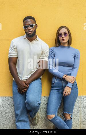 Photo verticale de deux personnes multiraciales élégantes dans des vêtements décontractés en denim et des lunettes de soleil appuyées sur un mur urbain Banque D'Images