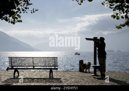 Femme sur le front de mer avec son chien près d'un banc et regardant avec un télescope au-dessus d'un lac majeur avec montagne et îles Brissago à Ascona Banque D'Images