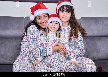 Trois filles sont assises sur un canapé, portant des pyjamas assortis et des chapeaux de père noël. Ils sourient et semblent apprécier la compagnie de l'autre Banque D'Images