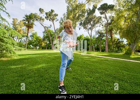 Homme latin en chemise blanche et Jean bleu dansant sur l'herbe d'un parc, mains à la caméra, mise au point sélective. Banque D'Images