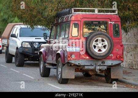 Un Land Rover Defender rouge avec porte-bagages de toit et roue de secours montés à l'arrière, stationné dans une rue de banlieue avec un véhicule utilitaire blanc visible devant. Banque D'Images