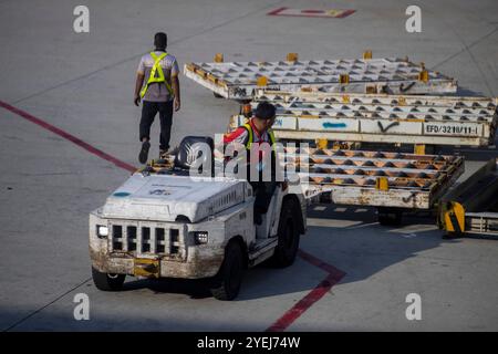 Membre de l'équipe au sol conduisant un véhicule de remorquage de bagages tandis qu'un autre travailleur marche à proximité sur le tarmac de l'aéroport, participant aux opérations de manutention de fret. Banque D'Images