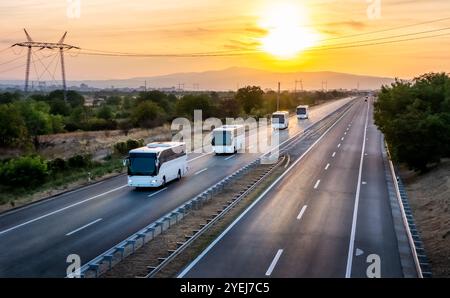 Série de quatre bus blancs modernes voyageant sur une large autoroute avec un beau coucher de soleil dramatique. Convoi de bus. Transport routier de passagers Banque D'Images