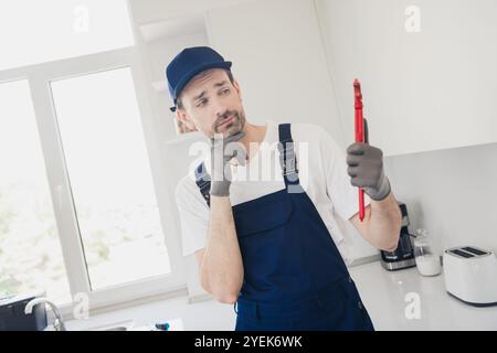 Jeune bricoleur réfléchi en salopette bleue contemplant une tâche de plomberie dans un environnement de cuisine moderne Banque D'Images