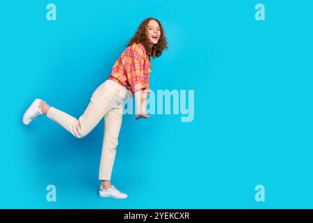 Jeune femme joyeuse avec les cheveux roux bouclés frappe une pose ludique dans une tenue décontractée vibrante sur un fond bleu vif Banque D'Images