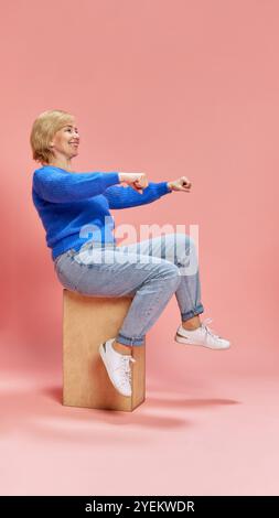 Femme âgée heureuse et souriante en jeans et pull bleu, assise sur la boîte et faisant semblant de rouler, conduisant une voiture sur fond de studio rose Banque D'Images