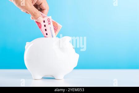 Main tenant un billet de 100 Yuan, le laissant tomber dans une tirelire blanche, avec la monnaie chinoise Yuan placée sur une table contre un bleu clair. Sauvegarde et gestion Banque D'Images