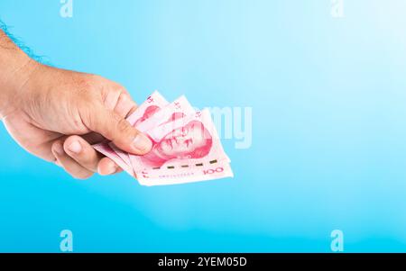 Main tenant un billet de 100 Yuan, le laissant tomber dans une tirelire blanche, avec la monnaie chinoise Yuan placée sur une table contre un bleu clair. Sauvegarde et gestion Banque D'Images