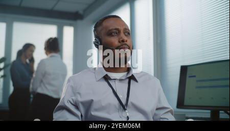 Bureau du centre d'appels : portrait rapproché d'un sympathique spécialiste de l'assistance technique afro-américaine en matière de micro-casques/oreillettes souriant, regardant la caméra. Équipe d'opérateurs de hotline non concentrée en arrière-plan. Banque D'Images