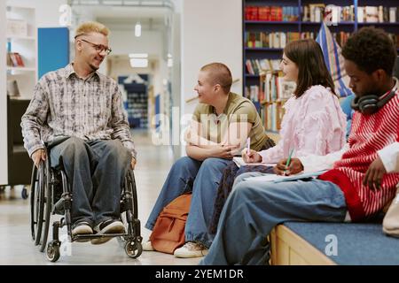 Groupe d'étudiants divers conversant dans un cadre de bibliothèque, y compris individuel en fauteuil roulant s'engageant avec des pairs entourés de livres et de coins salon confortables Banque D'Images