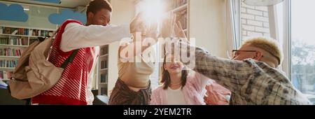 Groupe d'étudiants divers haut-fiving et souriant à l'intérieur de la bibliothèque remplie d'étagères, célébrant leur succès sur le projet. Lumière du soleil éclairant à travers les fenêtres, créant une ambiance joyeuse Banque D'Images