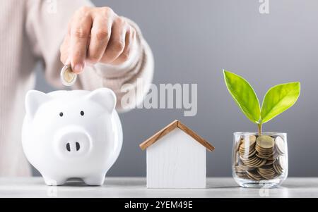 L'homme laisse tomber des pièces de monnaie dans une tirelire blanche, un pot en verre de pièce de monnaie et un modèle de maison en bois à côté de lui. Souligne le concept d'économiser de l'argent pour la future maison Banque D'Images