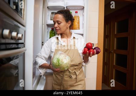 Une femme dans un tablier de cuisine sélectionne des légumes frais dans un réfrigérateur, se préparant pour un repas. L'image véhicule un mode de vie accueillant et sain, empha Banque D'Images