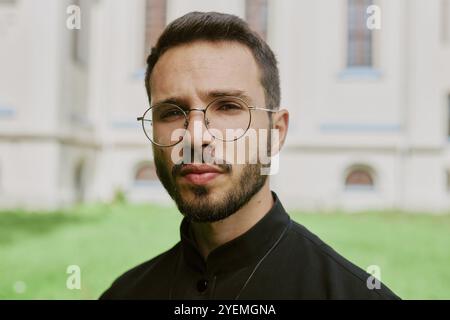 Portrait moyen en gros plan du beau jeune prêtre caucasien avec verre rond habillé en robe noire pour la photo Banque D'Images