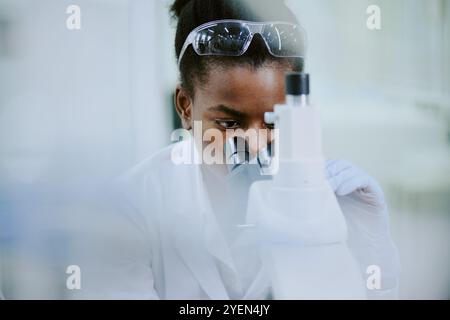 Scientifique afro-américaine travaillant en laboratoire se concentrant sur l'examen microscopique portant une blouse de laboratoire et des lunettes de sécurité analysant soigneusement les résultats avec une concentration précise Banque D'Images