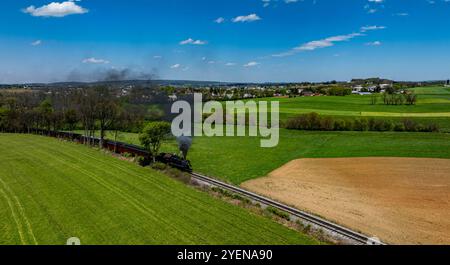 Une locomotive à vapeur souffle des nuages de fumée noire tout en voyageant à travers des champs verts vibrants sous un ciel bleu vif. Le paysage rural paisible Banque D'Images