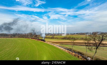 Un train à vapeur s'élance le long d'une piste qui traverse des champs verdoyants, avec une fumée bouillonnante contrastant avec le ciel bleu clair. Le Landsca rural Banque D'Images