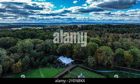 La photographie capture une vue à vol d'oiseau d'une forêt tentaculaire ornée d'un feuillage d'automne brillant. Une tente événementielle est visible, nichée parmi les arbres, sous un ciel dramatique rempli de nuages. Banque D'Images