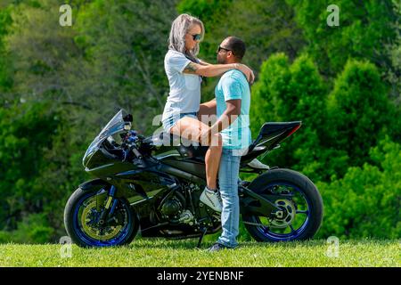 Un couple multiracial pose avec amour à côté de leur moto dans un champ verdoyant et luxuriant. Embrassés, ils sourient avec fierté, capturant à la fois leur lien profond A. Banque D'Images