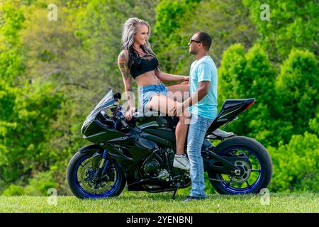 Un couple multiracial pose avec amour à côté de leur moto dans un champ verdoyant et luxuriant. Embrassés, ils sourient avec fierté, capturant à la fois leur lien profond A. Banque D'Images