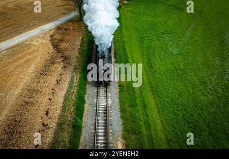 Une locomotive à vapeur d'époque souffle de la fumée blanche alors qu'elle se déplace le long des voies ferrées entourées de champs verdoyants et d'un chemin de gravier. Le ciel bleu clair ajoute à l'atmosphère rurale pittoresque. Banque D'Images