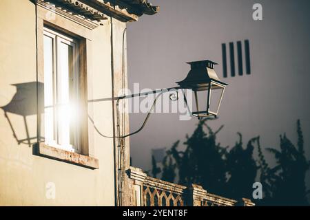 La lumière chaude du soleil se reflète sur une lanterne murale de style vintage montée sur le côté d'un ancien bâtiment en stuc avec une grande fenêtre. Ombres et créa de lumière douce Banque D'Images