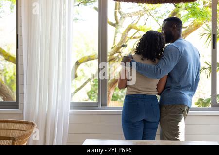 couple multiracial embrassant par fenêtre, profitant d'un moment paisible à la maison ensemble. Romance, amour, intime, détente, à l'intérieur, confortable Banque D'Images