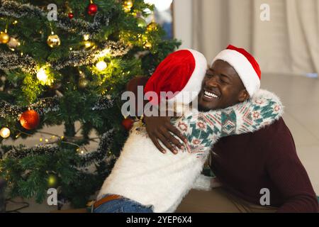 Couple multiracial souriant embrassé par l'arbre de Noël portant des chapeaux de Père Noël à la maison. Célébration, vacances, ensemble, festif, amour, bonheur Banque D'Images