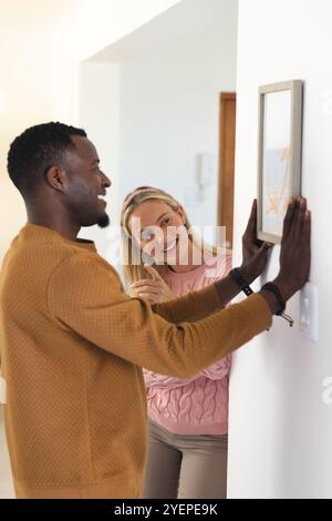 couple multiracial accrochant image à la maison, souriant et appréciant le moment. intérieur, collage, décoration, bonheur, travail d'équipe, compagnie Banque D'Images