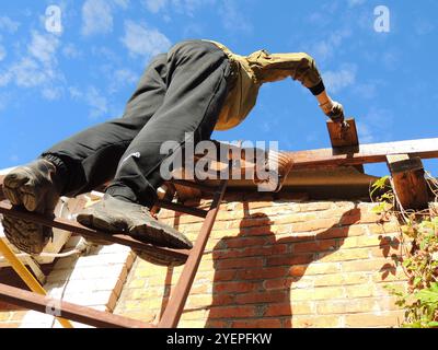 Un homme en vêtements de travail et gants debout sur une échelle métallique appuyée contre un mur de briques traite ou peint une structure de toit en bois Banque D'Images