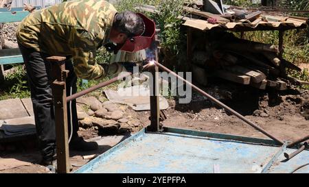 soudeur dans le casque de protection rouge et veste verte penchant sur la structure métallique dans la cour privée et travaillant sur le soudage des pièces en fer Banque D'Images