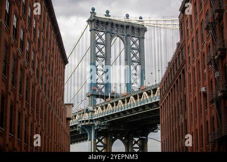 Cette image montre le pont de Manhattan encadré symétriquement par des bâtiments en briques rouges à DUMBO, Brooklyn, sous un ciel nuageux. Banque D'Images