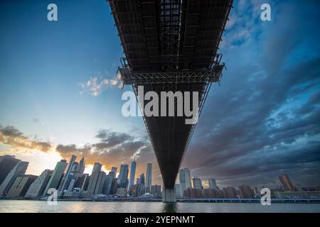 Photographie prise directement sous un pont, probablement le pont de Brooklyn, avec une vue sur les gratte-ciel de Manhattan au coucher du soleil. Banque D'Images