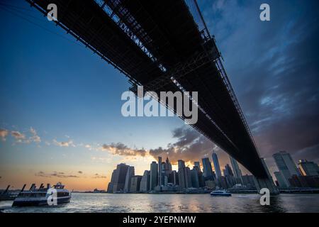 Photographie prise sous un pont, probablement le pont de Brooklyn, avec une vue sur les gratte-ciel de Manhattan au coucher du soleil. Banque D'Images