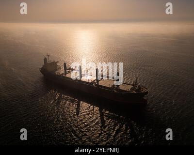 Drone aérien vidéo d'un cargo naviguant sur des eaux brumeuses avec des reflets de soleil, créant une scène maritime calme et mystérieuse, parfaite pour Banque D'Images