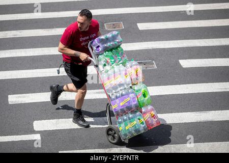 Un homme en chemise rouge pousse un camion à main chargé de plusieurs paquets de bouteilles de soda à travers un passage de ville. Banque D'Images
