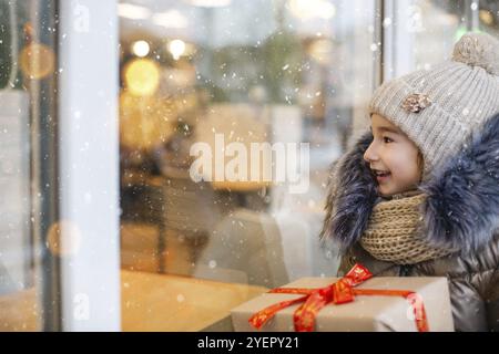 Portrait de fille joyeuse avec une boîte cadeau pour Noël près de la fenêtre de la boutique de verre en hiver avec la neige sur le marché de fête avec des décorations et des lumières. WA Banque D'Images