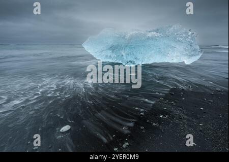 Un iceberg bleu chatoyant est emporté par les vagues sur une plage sombre, Diamond Beach, le lagon glaciaire de Joekulsarlon ou Joekulsarlon, dans le sud de l'Islande, en Islande Banque D'Images