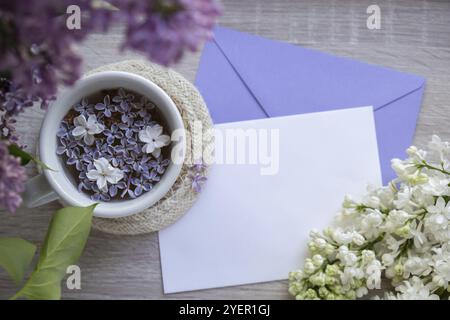 Lettre de papier d'espace de copie d'enveloppe vide avec thé noir savoureux dans une tasse blanche sur le rebord de la fenêtre avec des fleurs de lilas aromatiques. Composition printanière tasse de thé lilas Banque D'Images