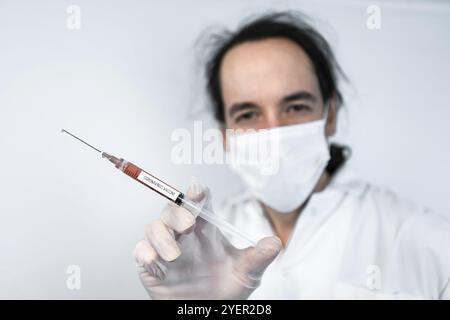 Plan de mise au point sélective d'une seringue avec un vaccin anti-coronavirus marqué au liquide rouge, tenu par la main gantée d'un scientifique masqué non focalisé au-dessus d'une grotte blanche Banque D'Images