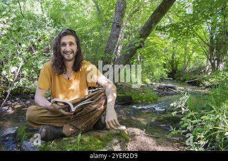 Jeune homme avec de longs cheveux assis les jambes croisées sur une pierre couverte de mousse sur le rivage d'une rivière dans les bois lisant un livre et souriant à la caméra Banque D'Images