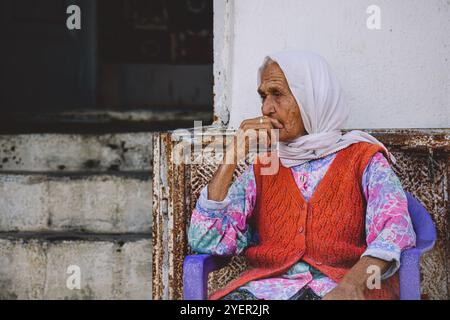 Une femme âgée portant une écharpe à motifs et un gilet vibrant se trouve pensivement devant sa maison en Cappadoce alors que le soleil se couche projetant une lumière chaude Banque D'Images
