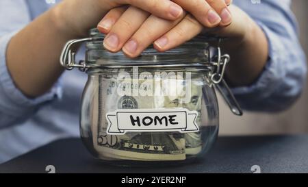 Mains féminines tenant pot en verre plein de billets de banque en dollars américains avec texte À LA MAISON. Préparation économisant de l'argent. Consommation modérée et econ Banque D'Images